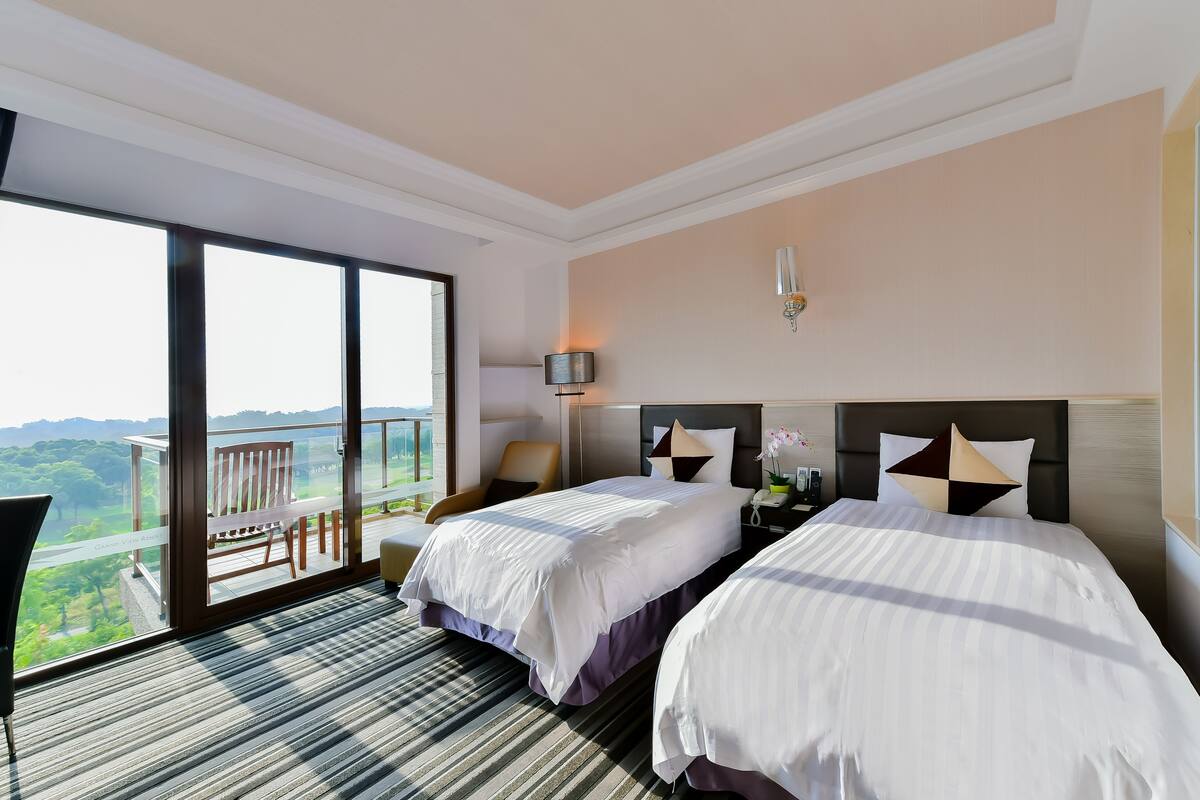 Habitación de hotel con dos camas, un ventanal amplio con vistas a zonas verdes, una silla y una mesita de noche con lámpara.
