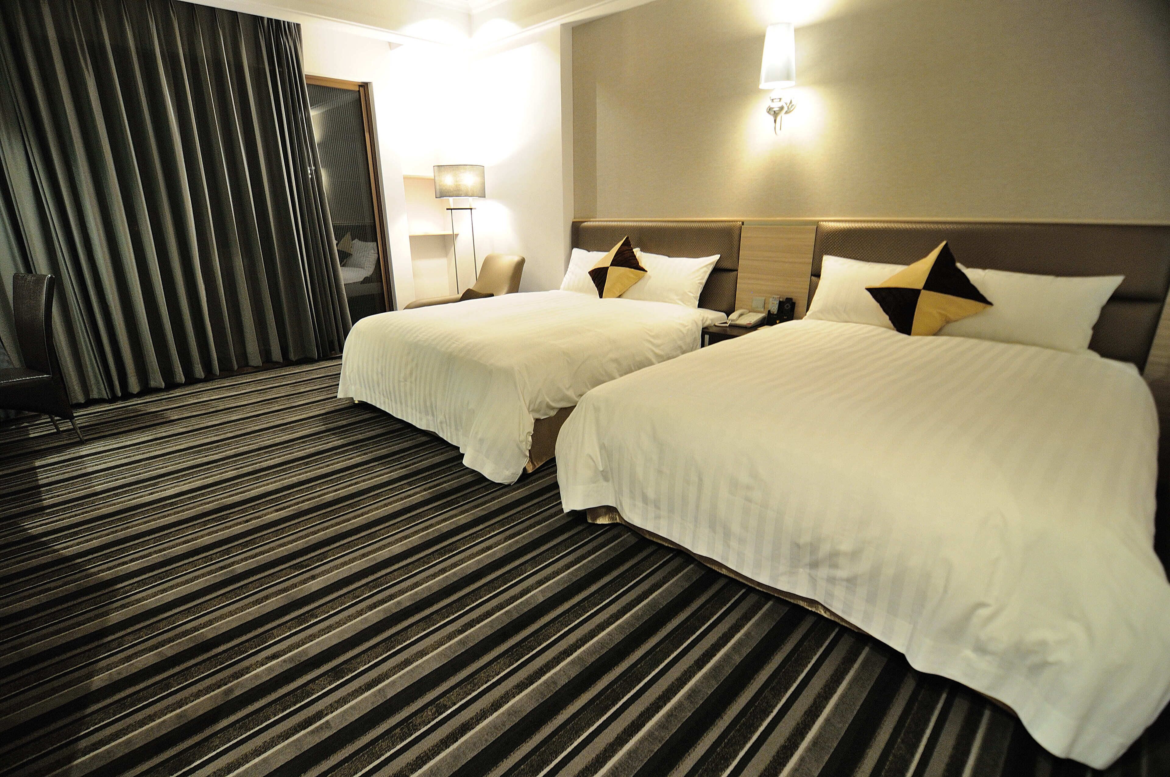 Una habitación de hotel con dos camas, un ventanal con cortinas, una mesita de noche con lámpara y suelo alfombrado.