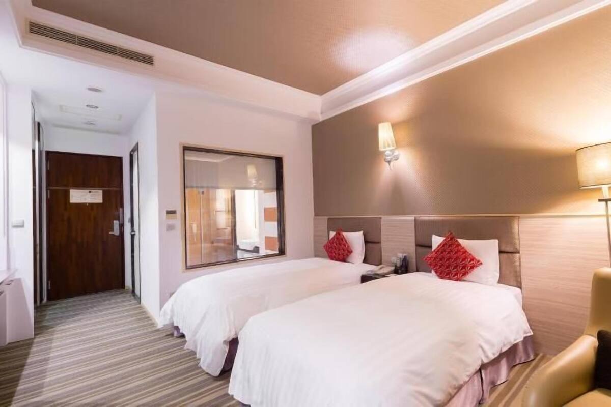 Habitación de hotel con dos camas, una zona de estar con una silla, un escritorio y una lámpara.