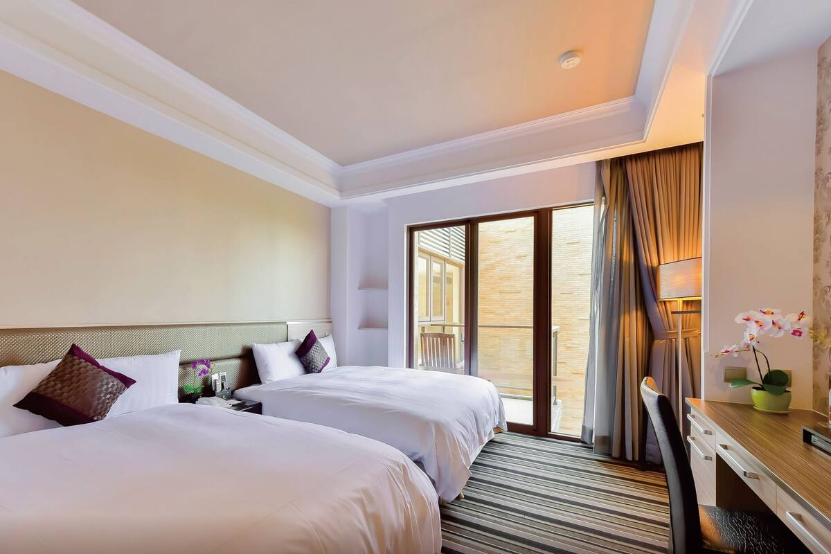 Habitación de hotel con dos camas, un escritorio, una silla, un ventanal con cortinas y vistas al exterior.