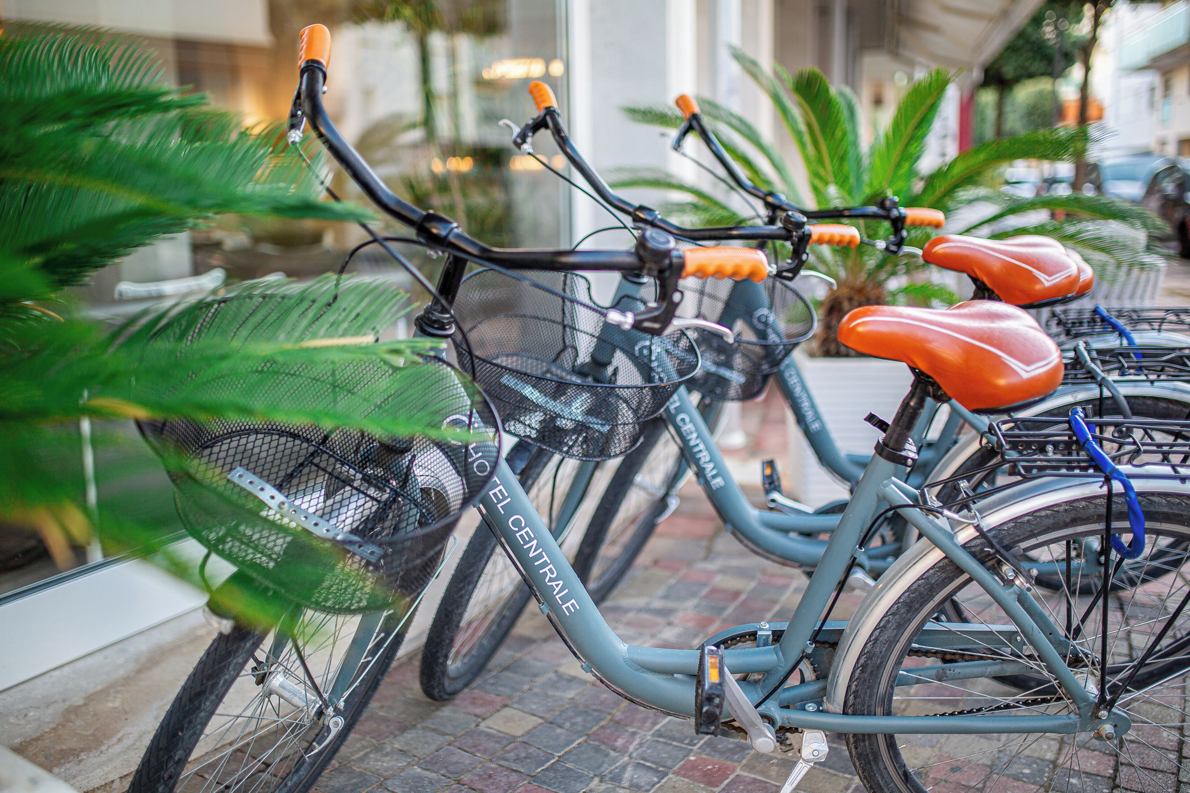 A row of bicycles with orange seats and a basket, parked on a brick pavement.