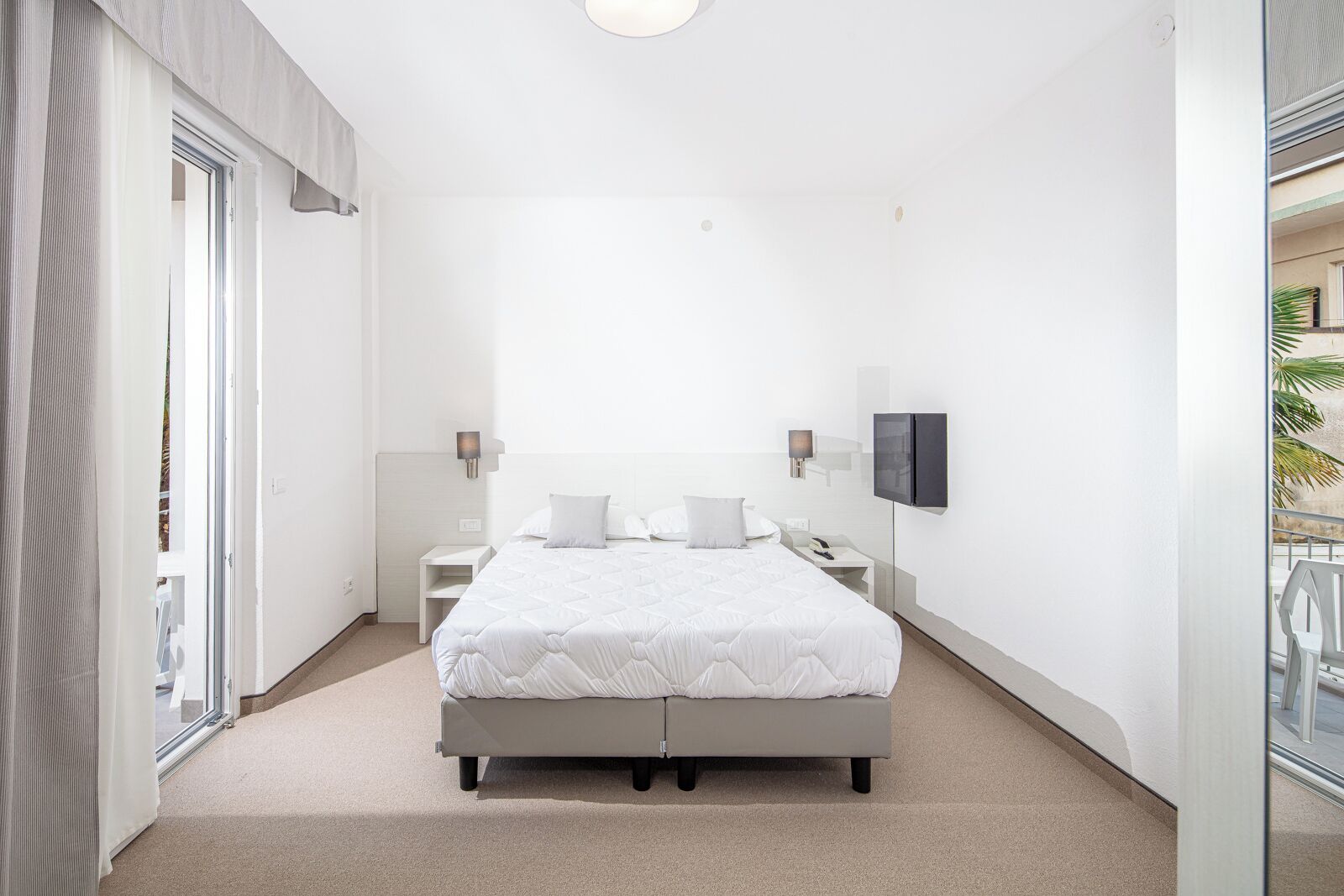 A modern hotel room with a large bed, a flat-screen TV, a bedside table, and a sliding glass door to the balcony.