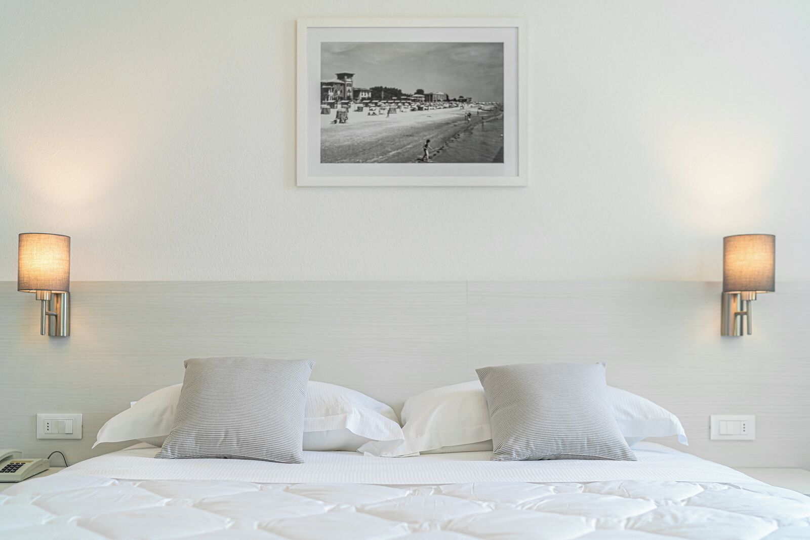 A hotel room with a bed, two pillows, two wall-mounted lamps, a framed photograph on the wall, and a telephone on the nightstand.