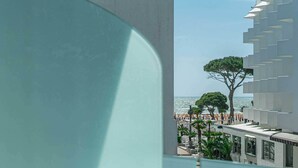 Vista desde un balcón sobre una playa, con un edificio moderno y un árbol.