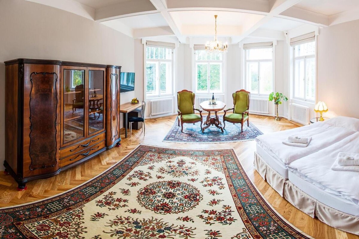 A hotel room with a large bed, a wooden cabinet, a dining table with chairs, and a chandelier.