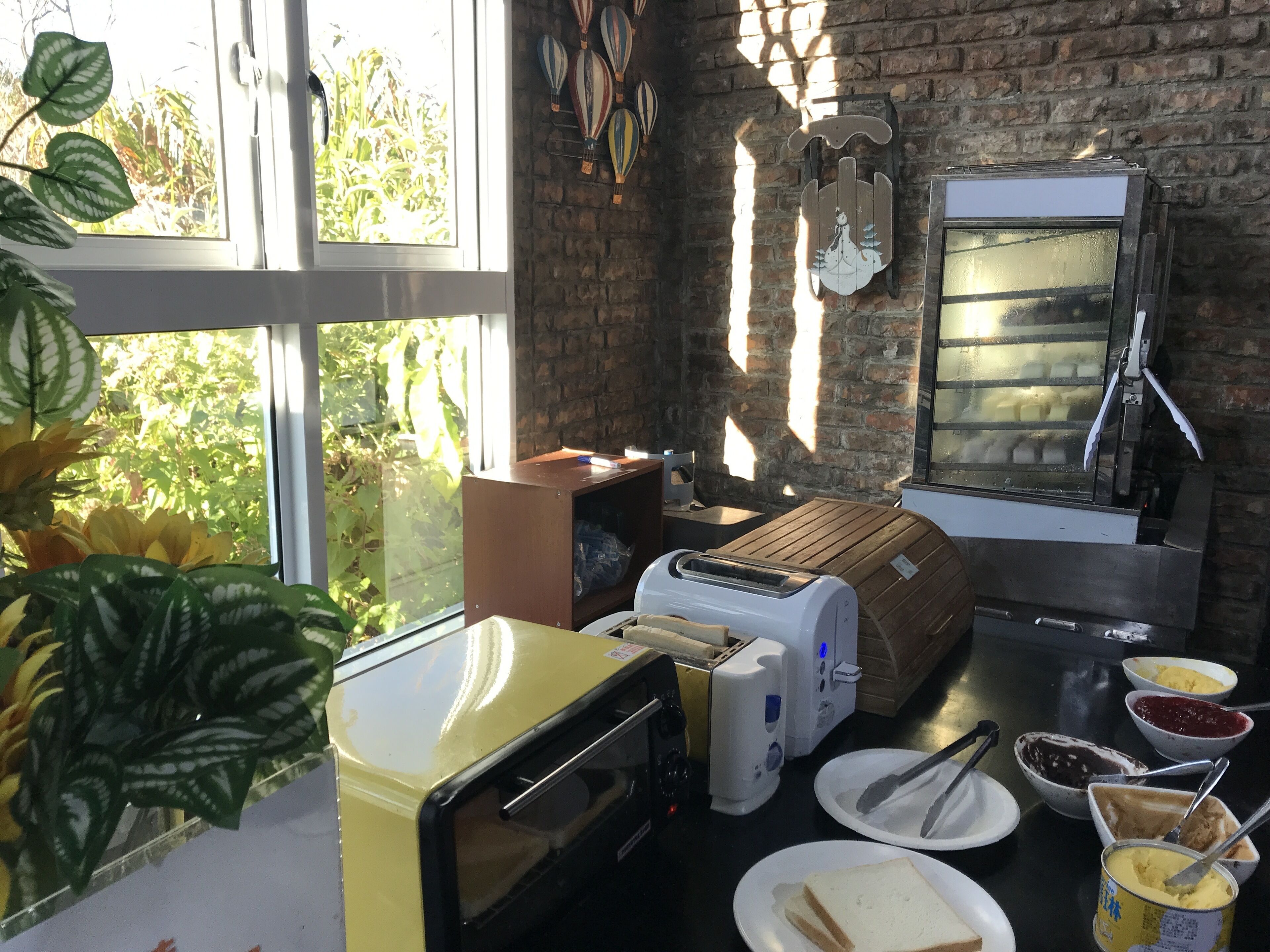 A kitchen with a brick wall, a window with plants, a toaster, a bread machine, and various food items.