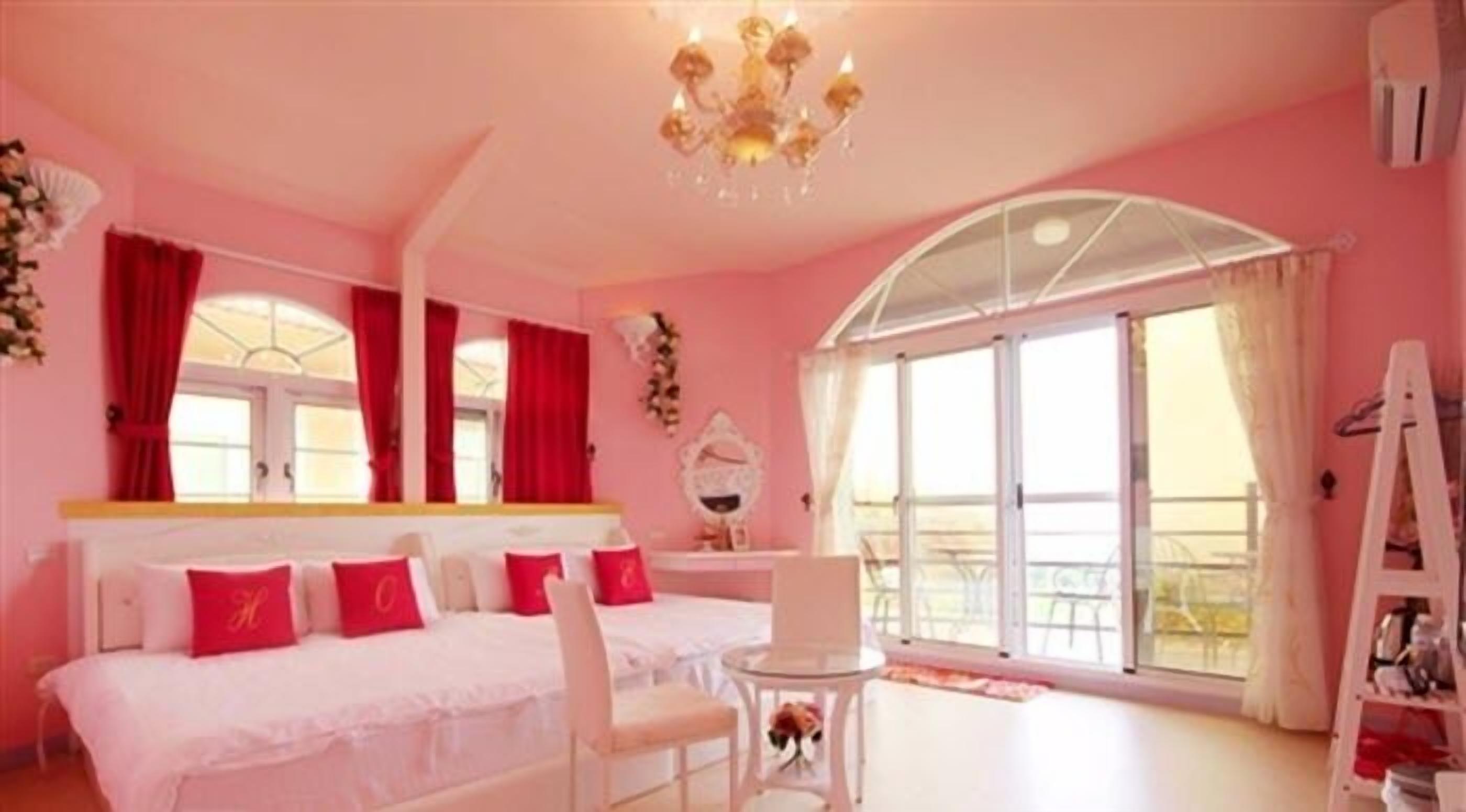 A pink-themed bedroom with two beds, a chandelier, a large window, and a white chair.