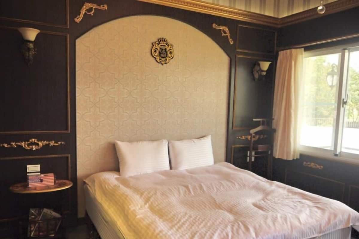 A hotel room with a large bed, a headboard with a crest, a small table with a lamp, and a window with curtains.