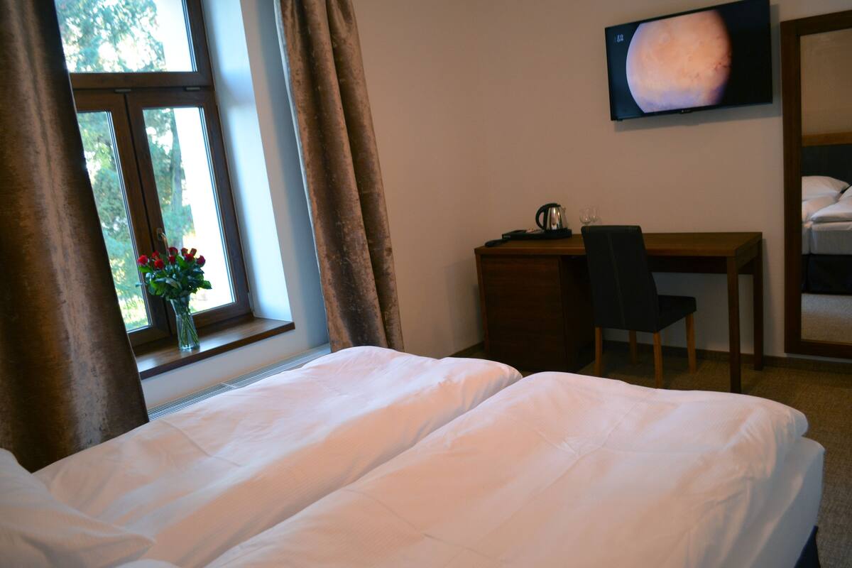 A hotel room with two beds, a desk with a chair, a TV on the wall, and a window with curtains.