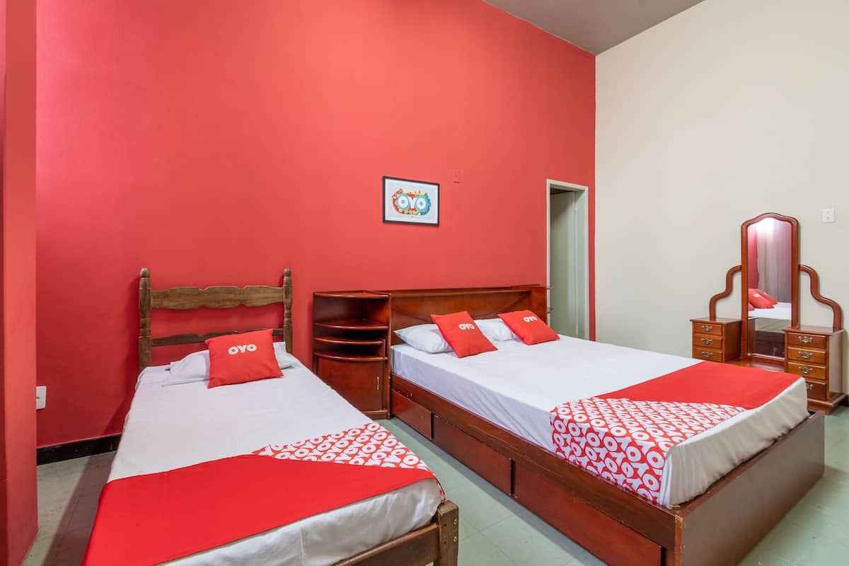 A room with two beds, each with a wooden headboard, red bedding, and a red pillow with 'OYO' branding. A wooden wardrobe and a mirror are also visible.