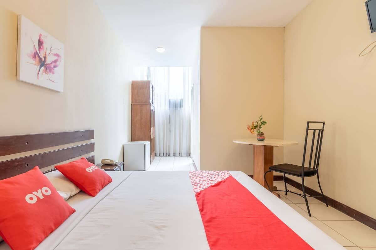 A hotel room with a bed, two red pillows, a bedside table with a vase, a chair, and a view of a corridor.