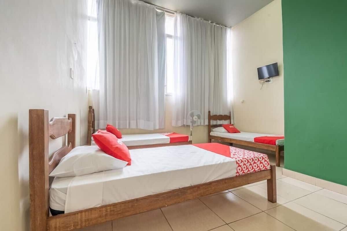 A room with three wooden beds, white bedding, red pillows, a green wall, a flat-screen TV, and a window with white curtains.