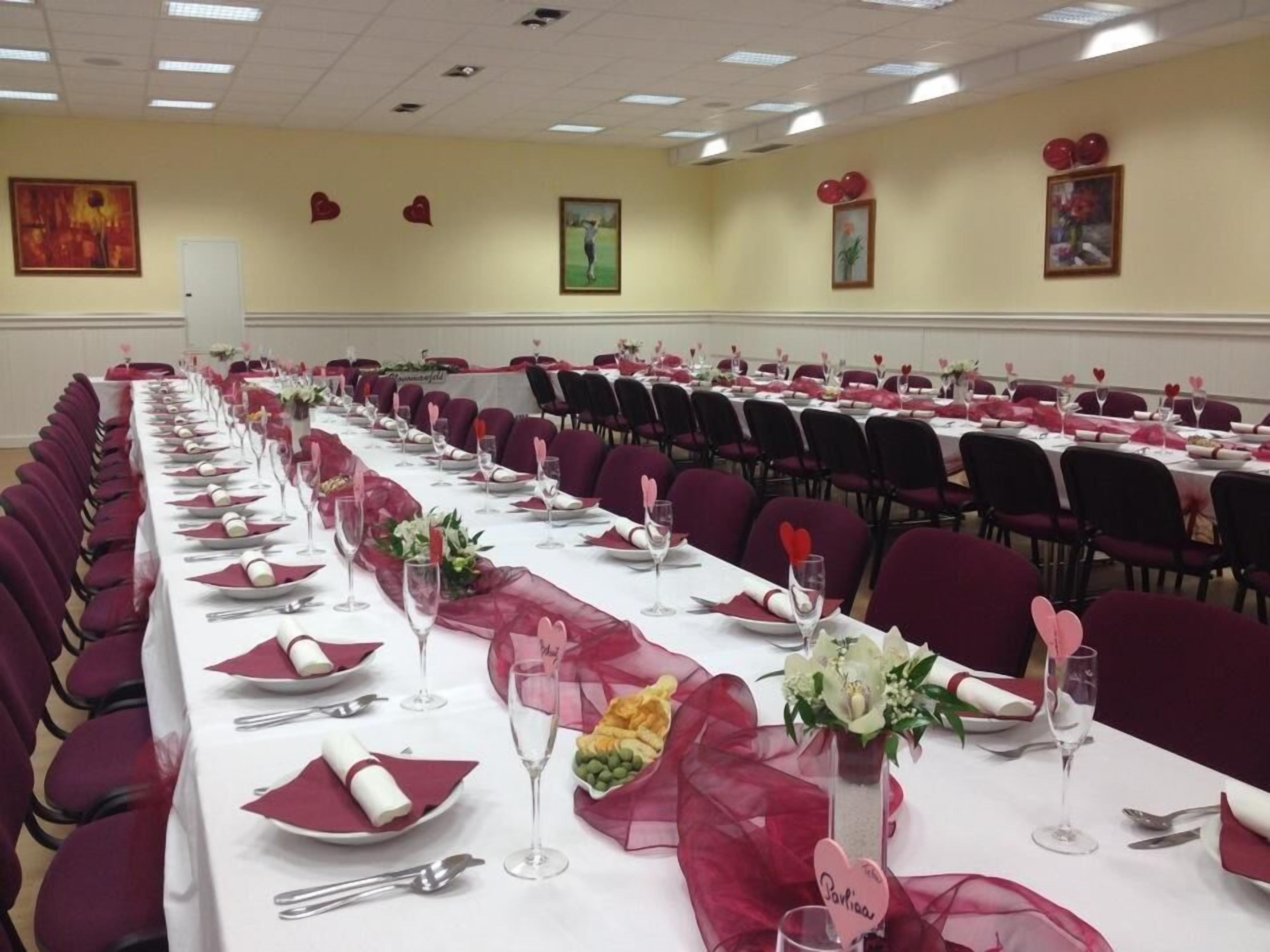 Uma sala de banquetes com longas mesas preparadas para um evento formal, com toalhas de mesa vermelhas e brancas, decorações em formato de coração e centros de mesa florais.