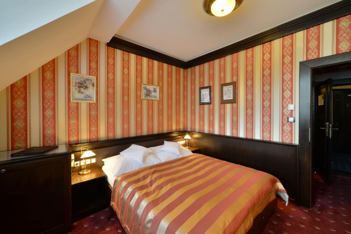 Ein Hotelzimmer mit Bett, Nachttischen und Schreibtisch. An der Wand hängen Bilder in Rahmen und rechts ist eine Tür zu sehen.