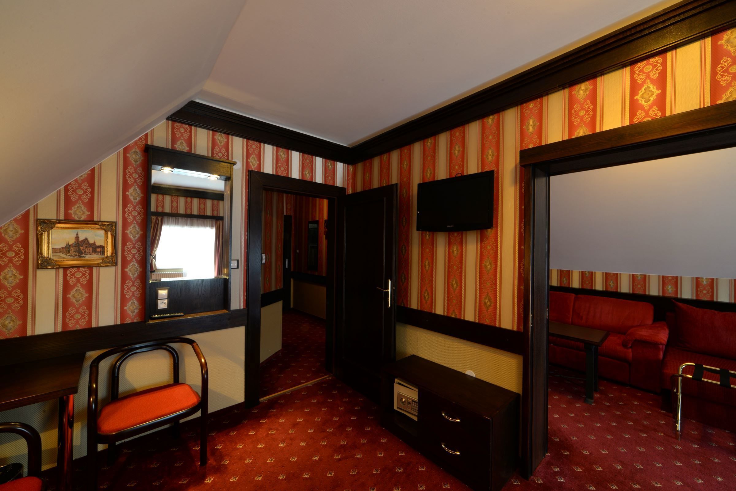 A hotel room with a red carpet, a sofa, a TV, a desk, and a chair.