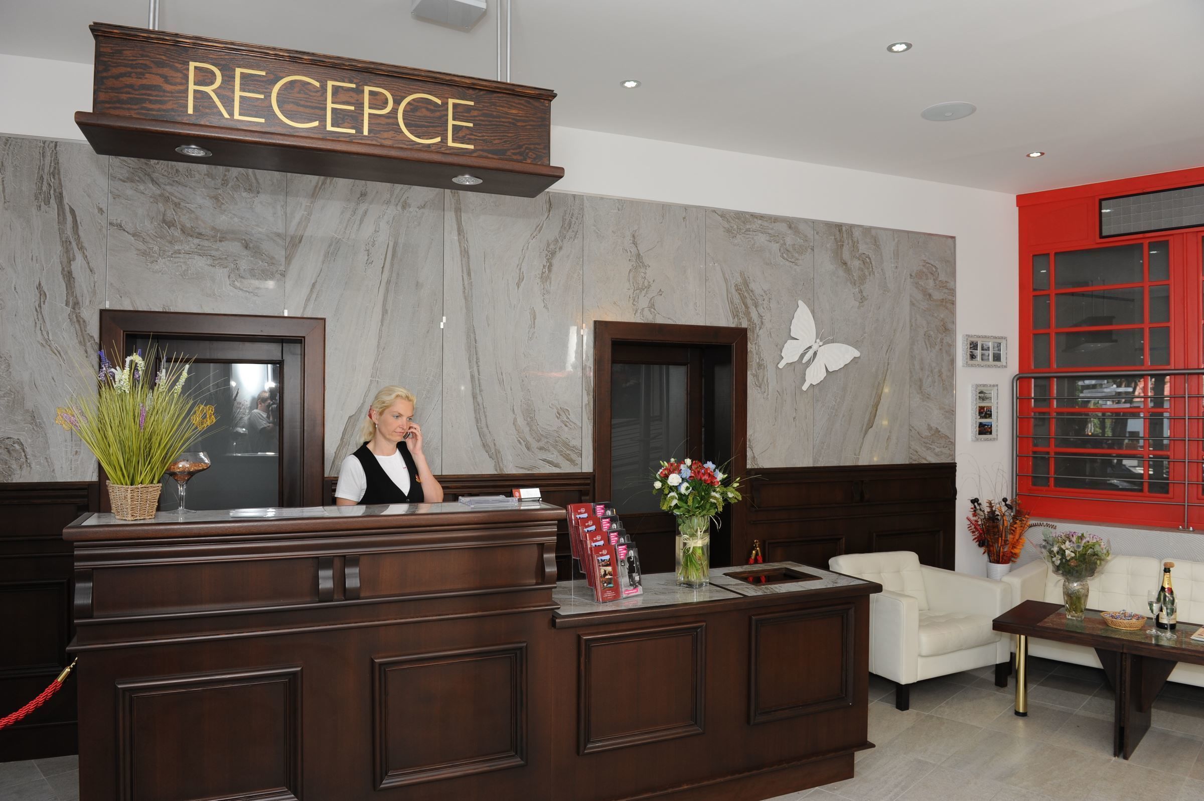 A reception desk with a sign reading "RECEPTION," a receptionist, a vase with flowers, and a red-framed window.