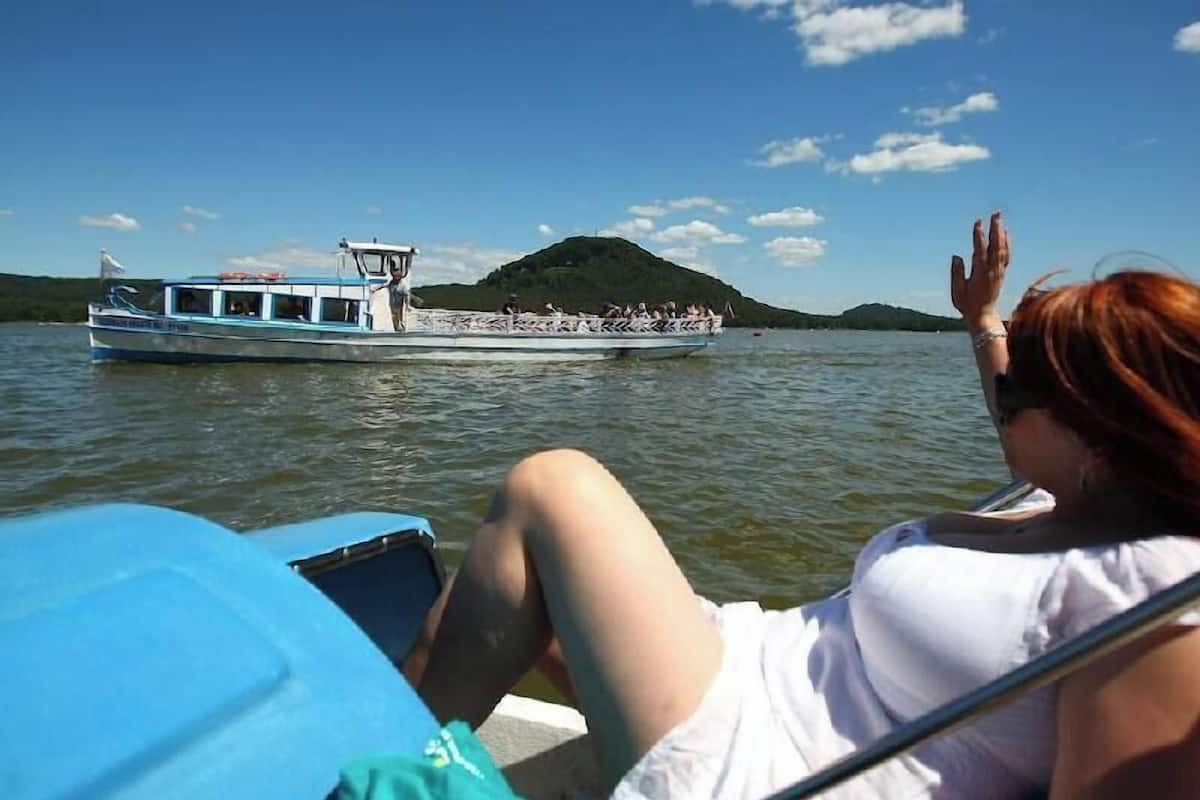 Ein Boot mit Passagieren auf ruhigem Gewässer, eine Person mit roten Haaren und ein grüner Hügel im Hintergrund.