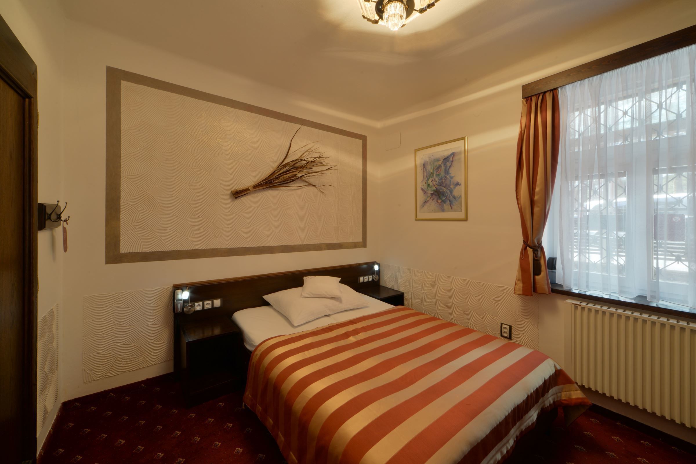 A hotel room with a bed, a nightstand, a wall-mounted lamp, a window with curtains, and a radiator.