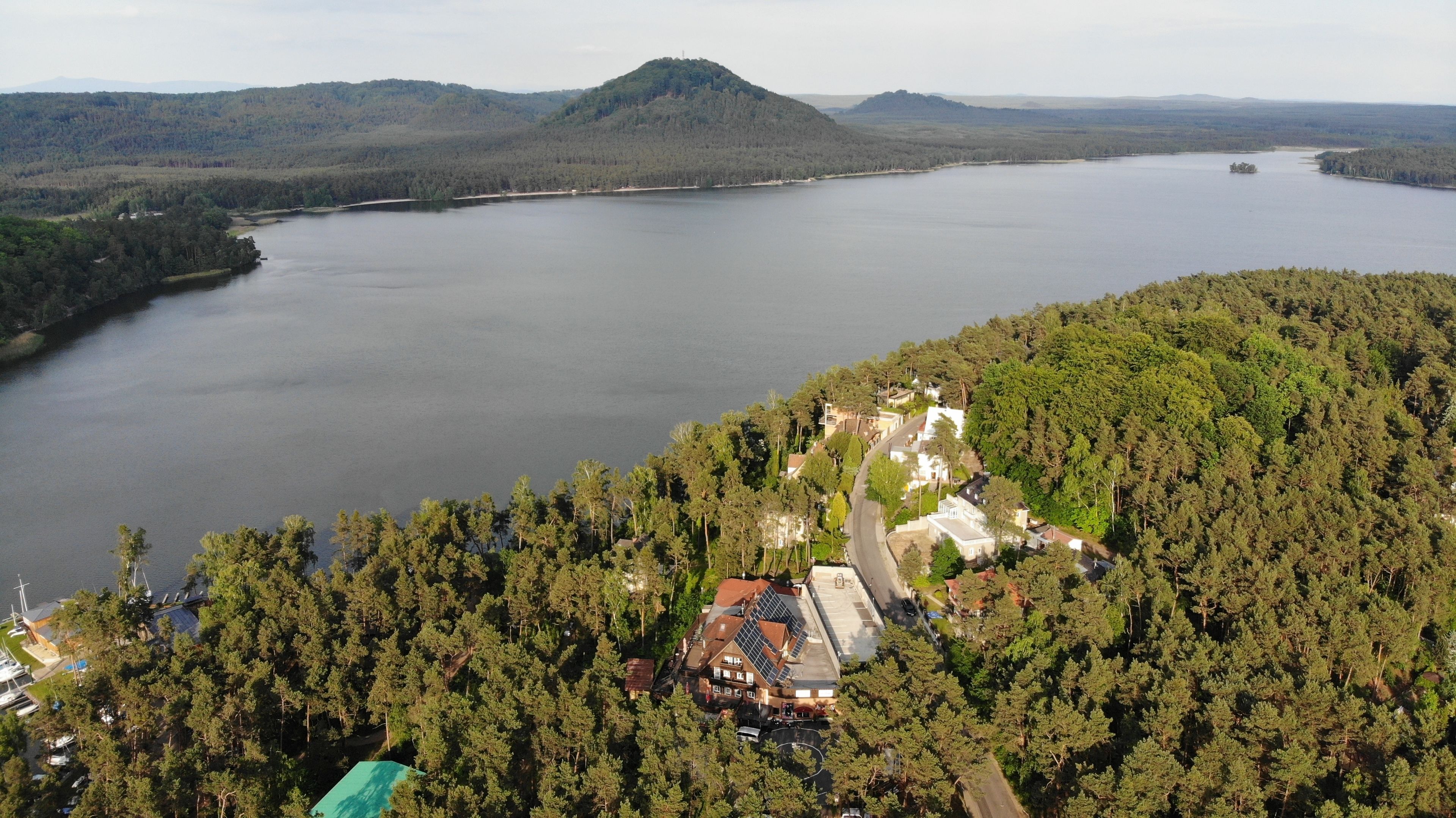 Una propiedad junto al lago con un camino que lleva a un edificio, rodeada de un bosque denso y colinas al fondo.