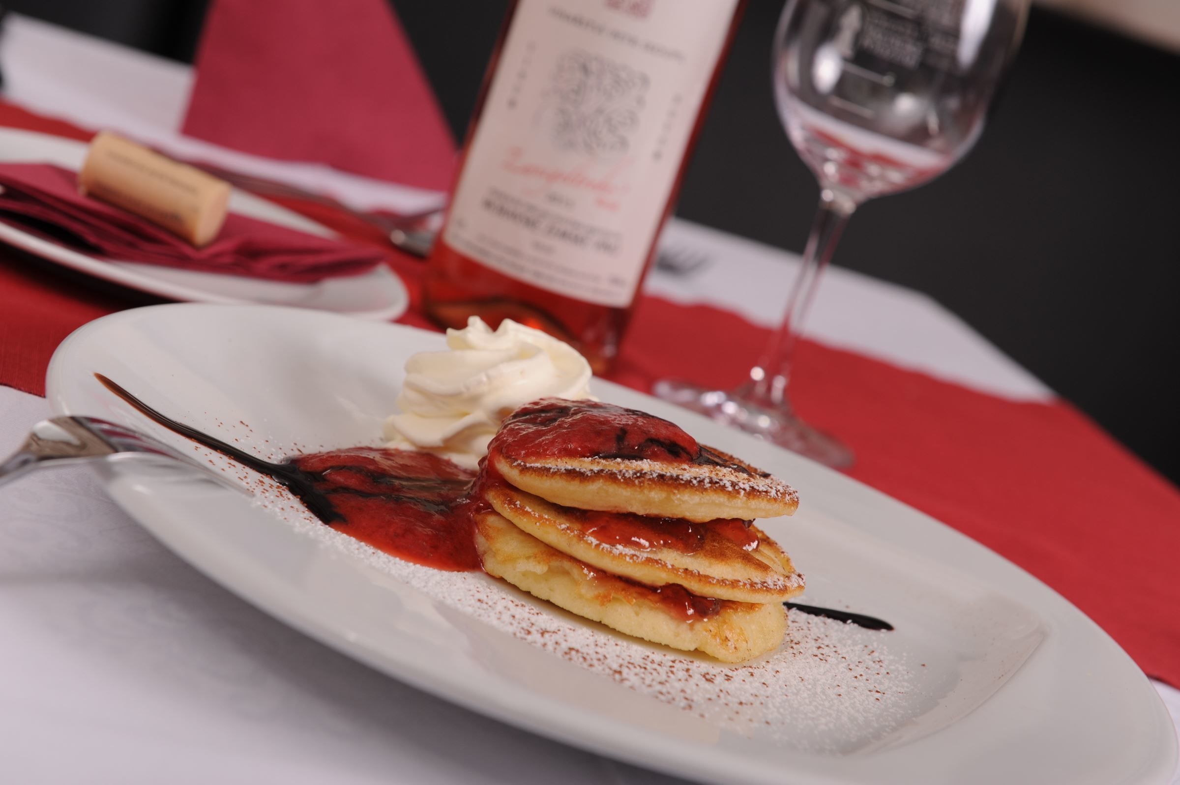 A dessert plate with pancakes, whipped cream, and a sauce, accompanied by a wine glass and a menu.