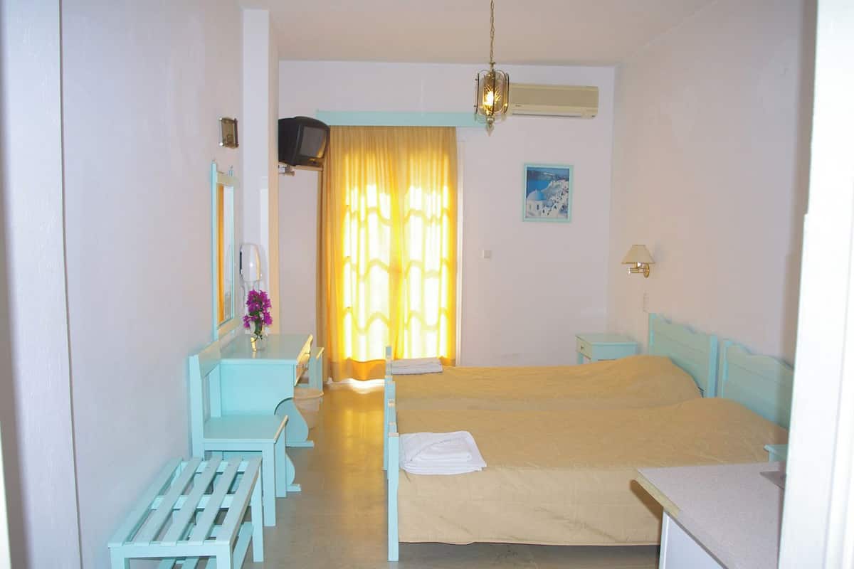 A room with a bed, a desk with a chair, a small table, a mirror, a television, and a window with curtains.