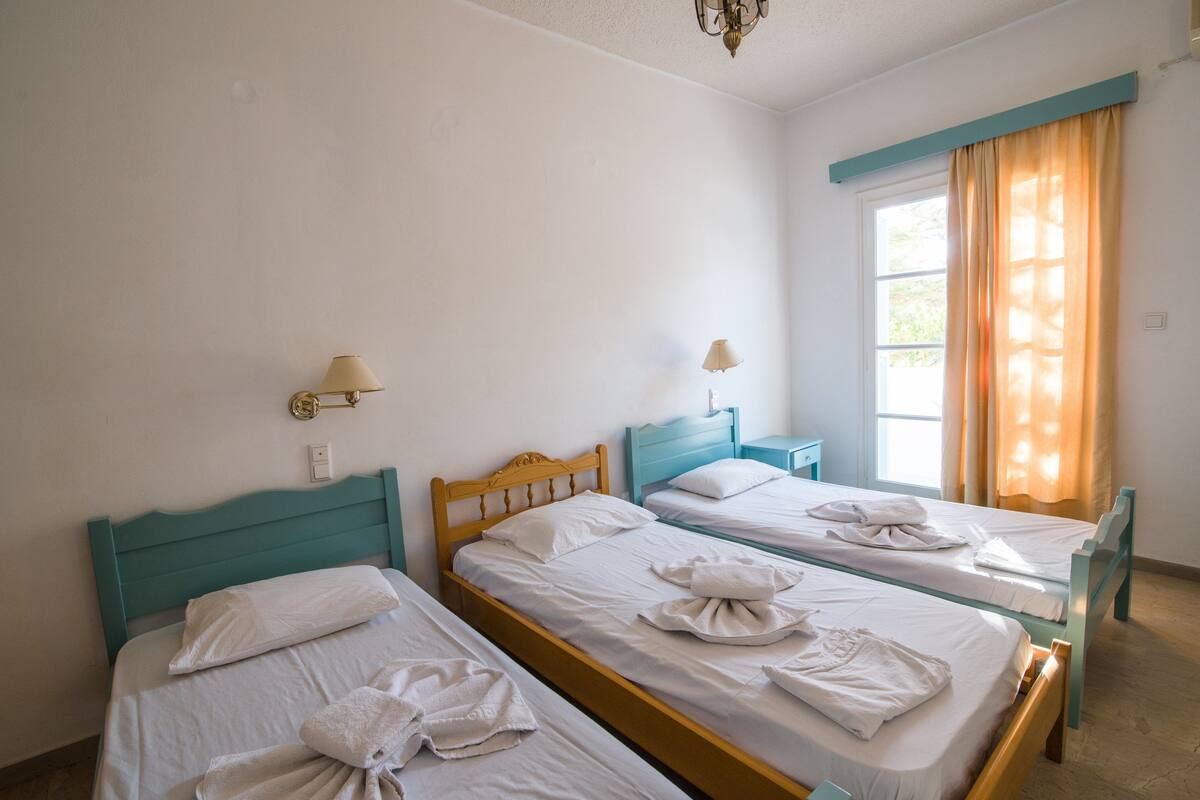 A hotel room with three single beds, each with a wooden headboard and white bedding. There are two bedside tables with lamps, and a window with curtains.