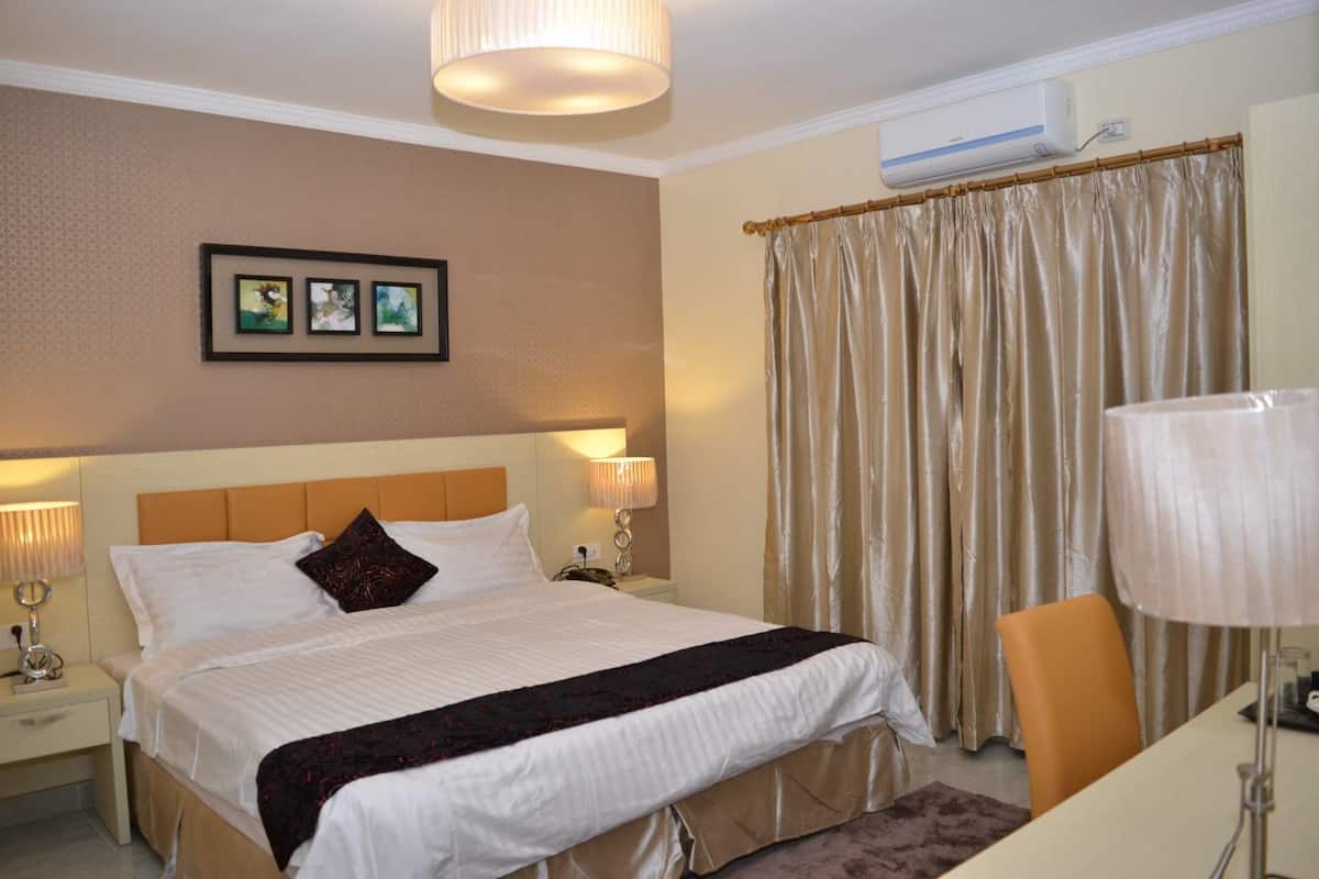 A hotel room with a large bed, two bedside lamps, a headboard, a framed picture on the wall, a window with curtains, and an air conditioning unit.