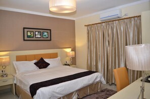 A hotel room with a large bed, two bedside lamps, a headboard, a framed picture on the wall, a window with curtains, and an air conditioning unit.