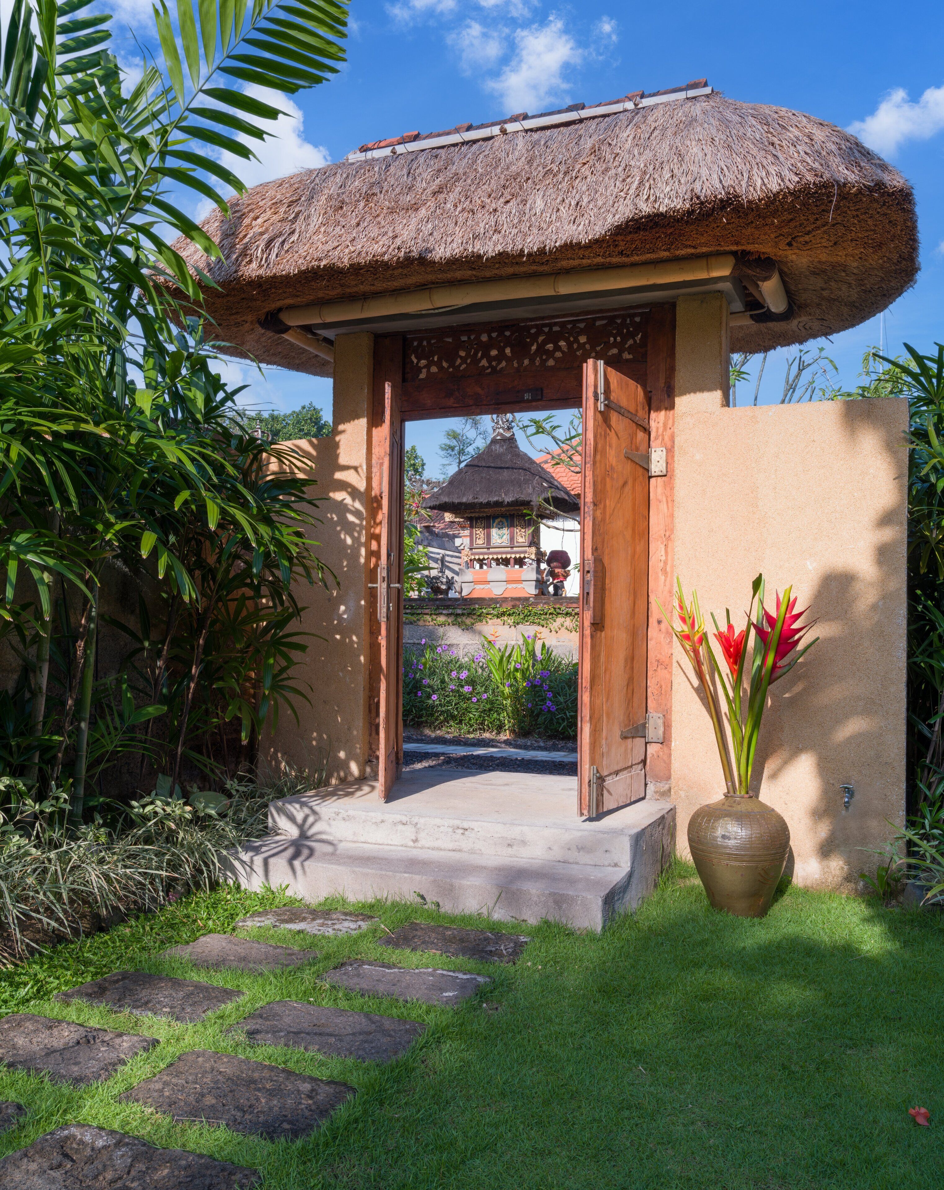 A thatched-roof structure with a wooden door, surrounded by greenery and a stone pathway leading to a building with a tiled roof.