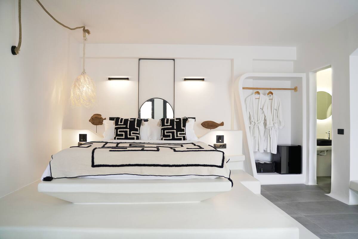 Un dormitorio moderno con una cama grande, mesitas de noche, una lámpara colgante, un espejo y un vestidor con batas colgadas.