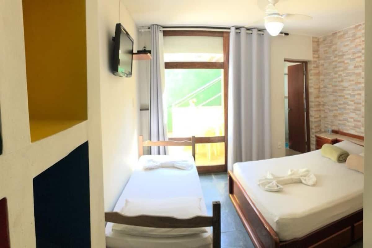 Quarto de hotel com duas camas, televisão, ventoinha no teto, janela com cortinas e parede com padrão de tijolos.