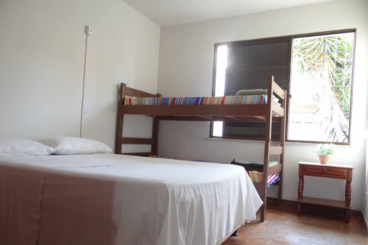Quarto com beliche, uma cama individual, uma mesinha com uma planta e janela com vista para o verde.