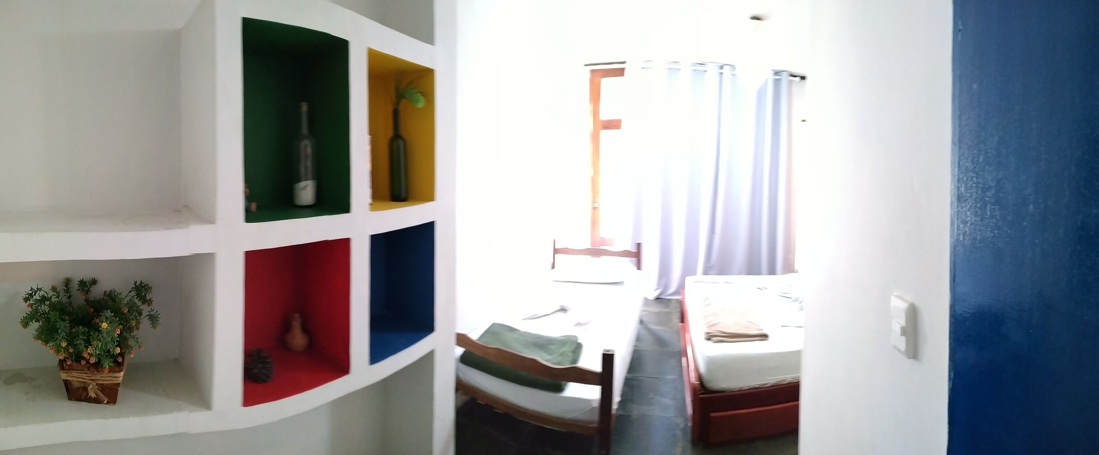 Una habitación con una cama, una silla, una estantería con objetos decorativos y una ventana con cortinas.
