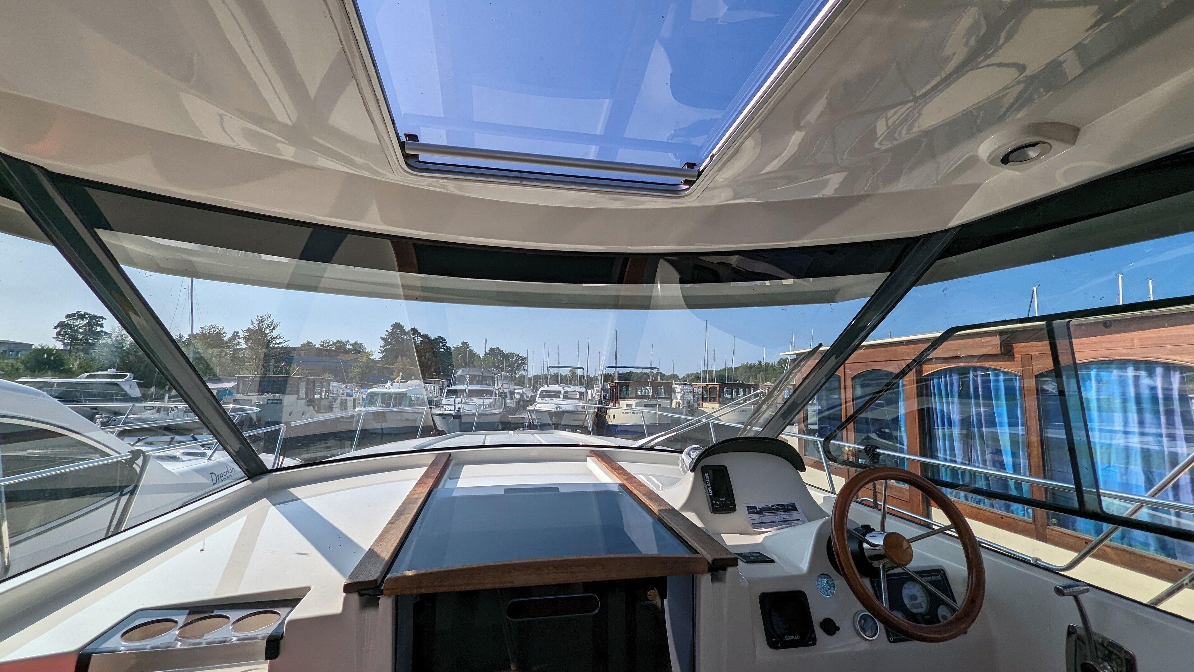 L'intérieur d'un bateau, avec un volant, un panneau de commande et une vue sur d'autres bateaux et les bâtiments du port de plaisance.