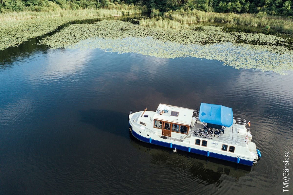 A boat with a covered deck and a cabin, floating on a calm body of water surrounded by greenery.