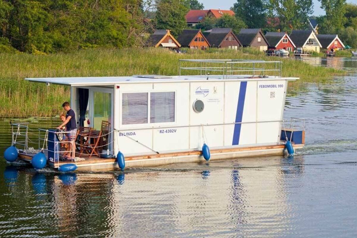 A houseboat with a covered deck, a small outdoor seating area, and a person standing at the entrance.