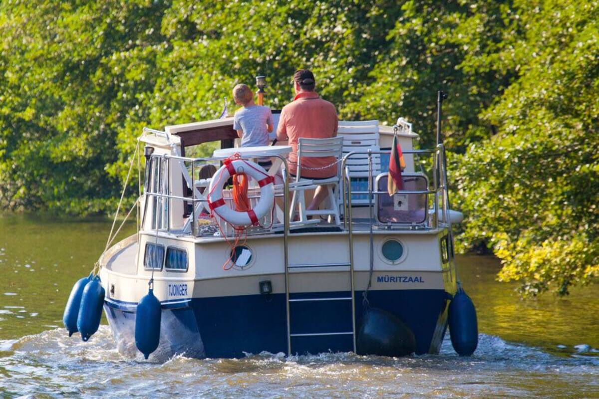 A boat with two people and a child, equipped with life jackets and a flag, moving on a river surrounded by greenery.