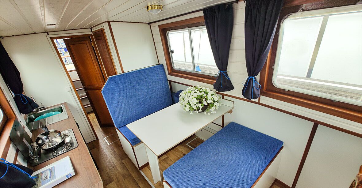 A compact cabin with a single table, a blue cushioned bench, a small window with curtains, and a wooden door.