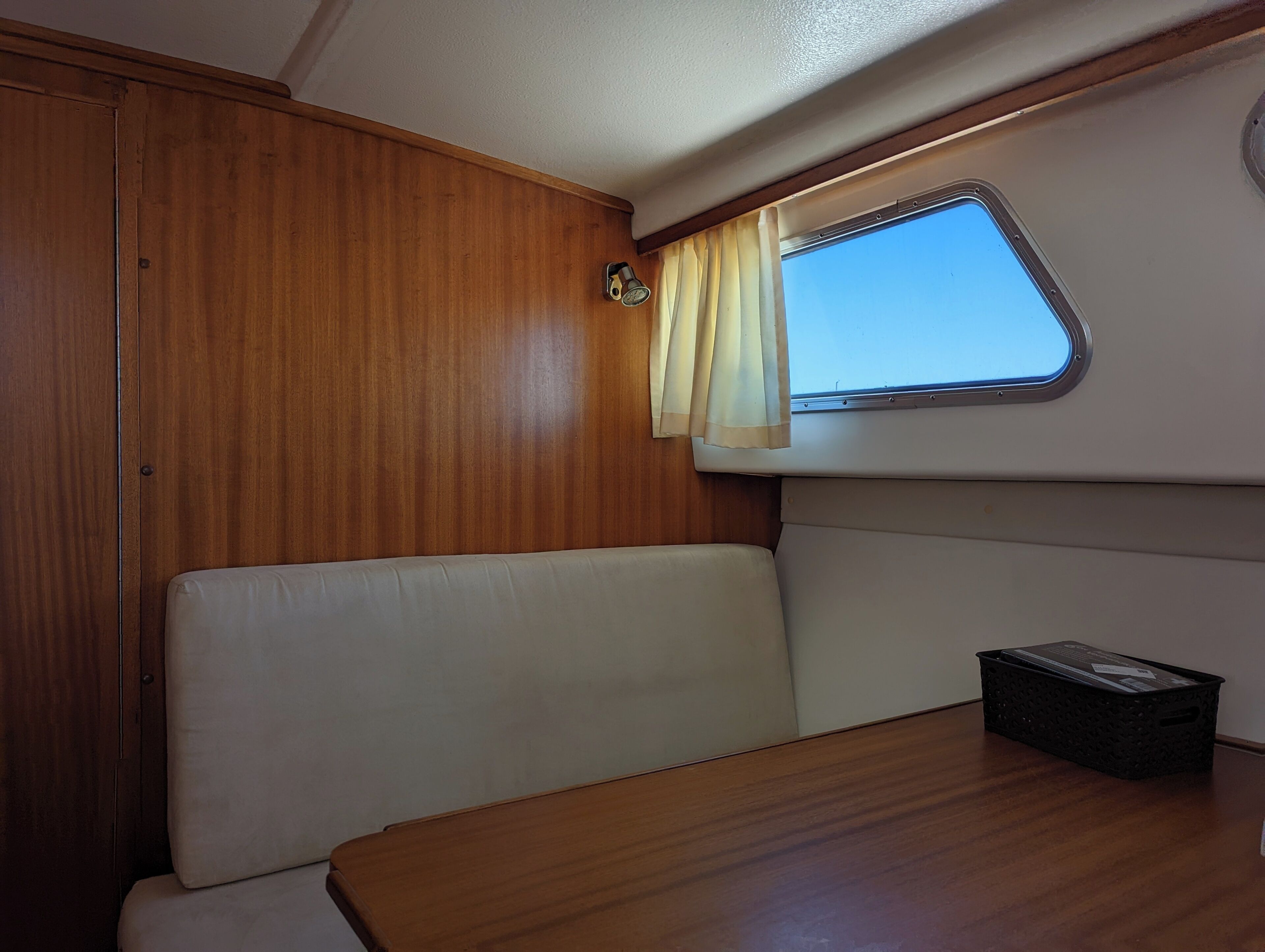 A compact, well-lit interior space with a wooden panel wall, a cushioned bench, a small table, and a window with curtains.