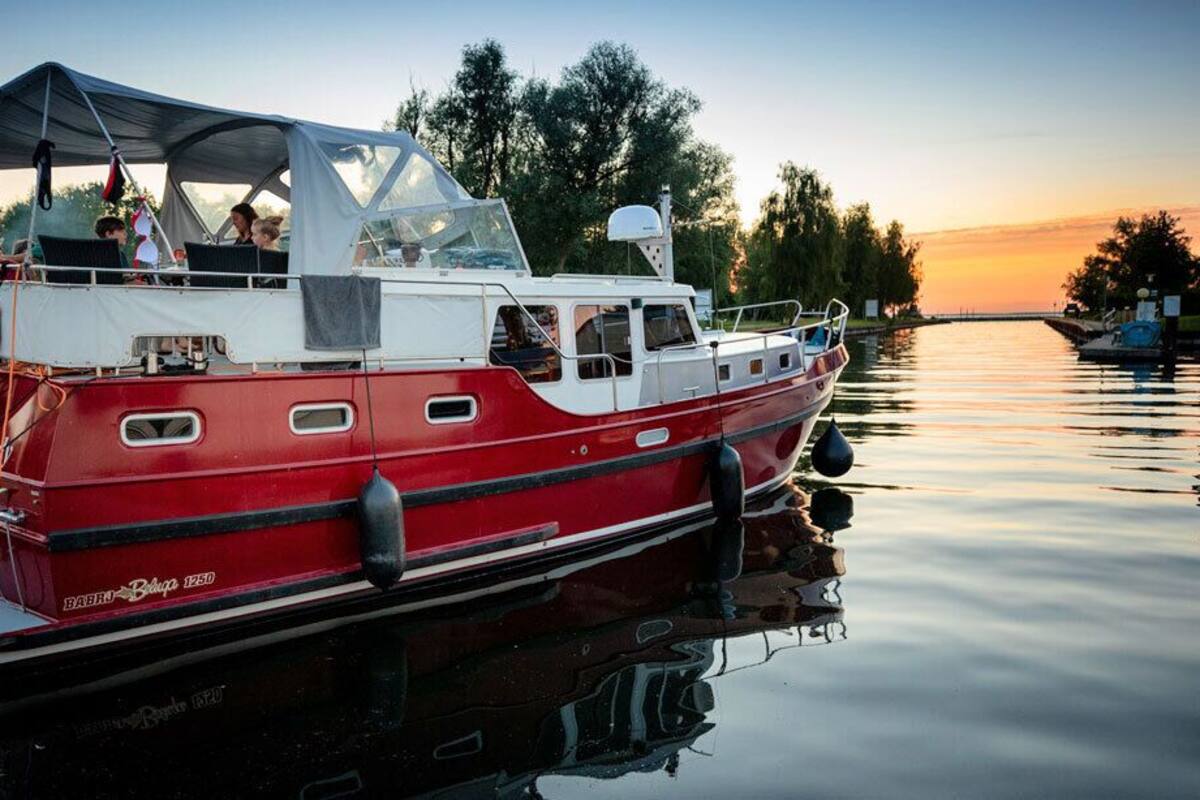 A red and white boat docked at a pier with a canopy, calm water, and trees in the background.