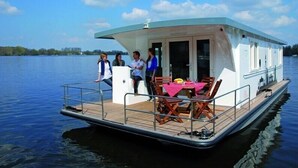 Una casa flotante con cubierta, un comedor con mesa y sillas, y un pequeño espacio de estar al aire libre.