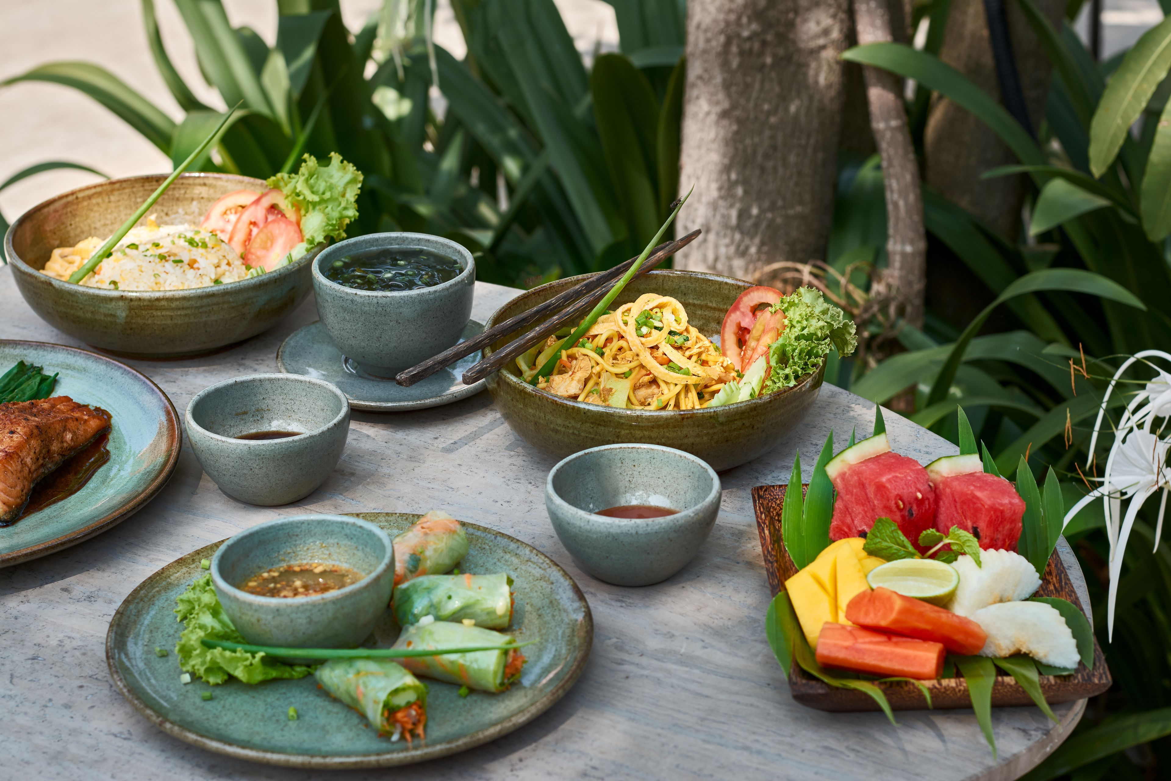 A table with various dishes including rice, noodles, and fresh vegetables, suggesting a meal setting.
