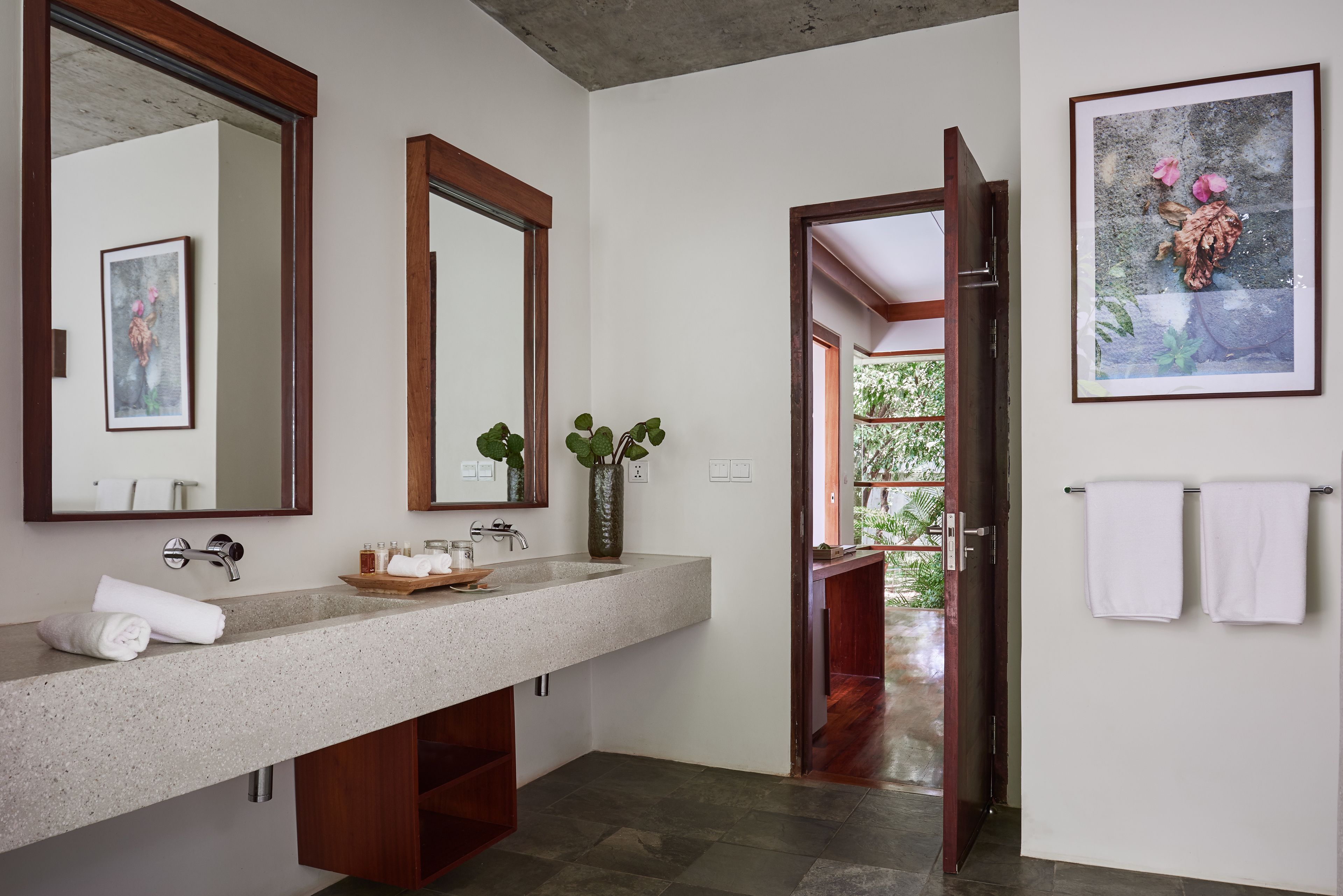 A modern bathroom with a large granite sink, two mirrors, a vase with green plants, and a framed picture on the wall.