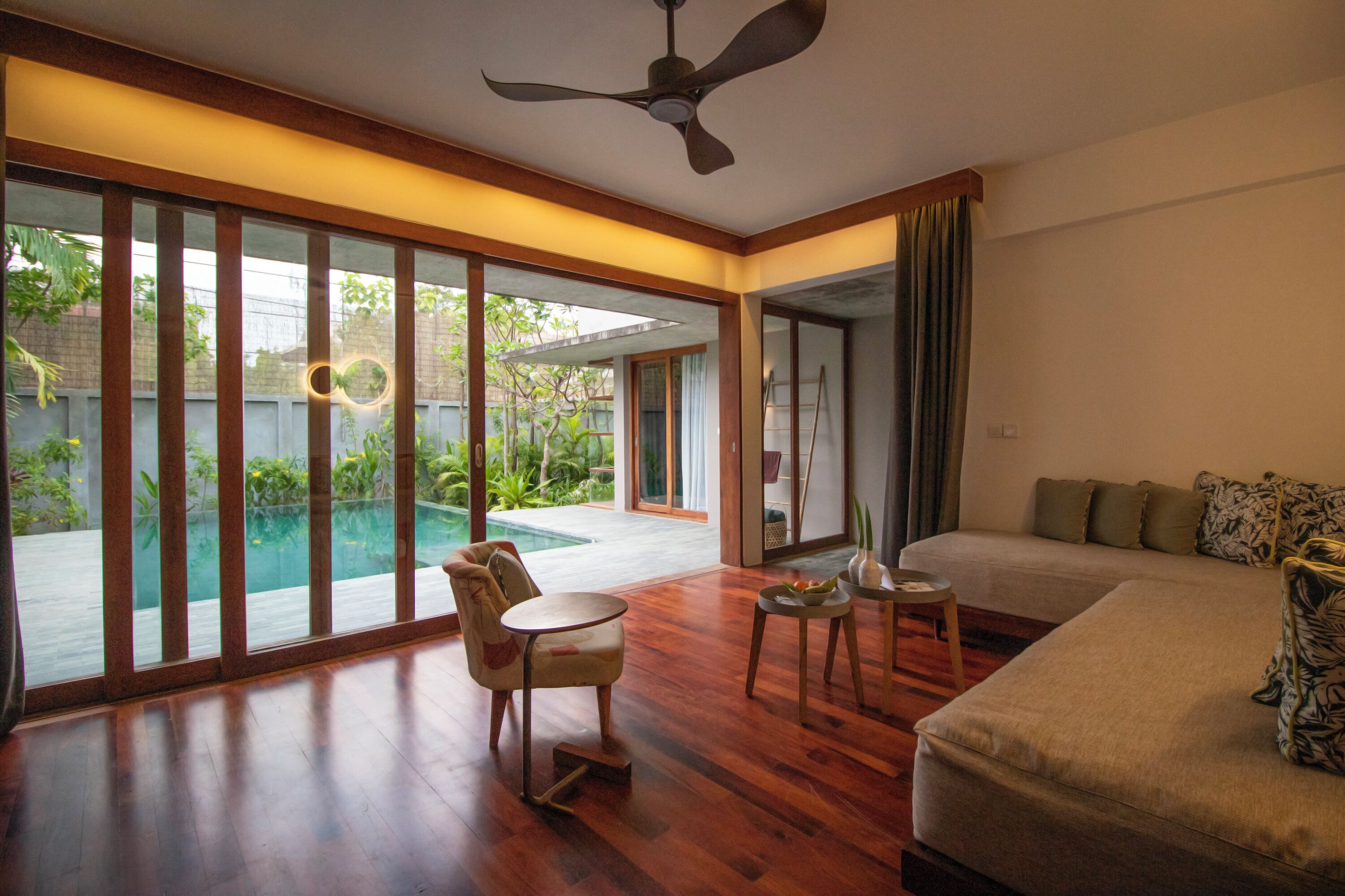 A spacious room with a large bed, a ceiling fan, a small table, and a view of a swimming pool and garden.
