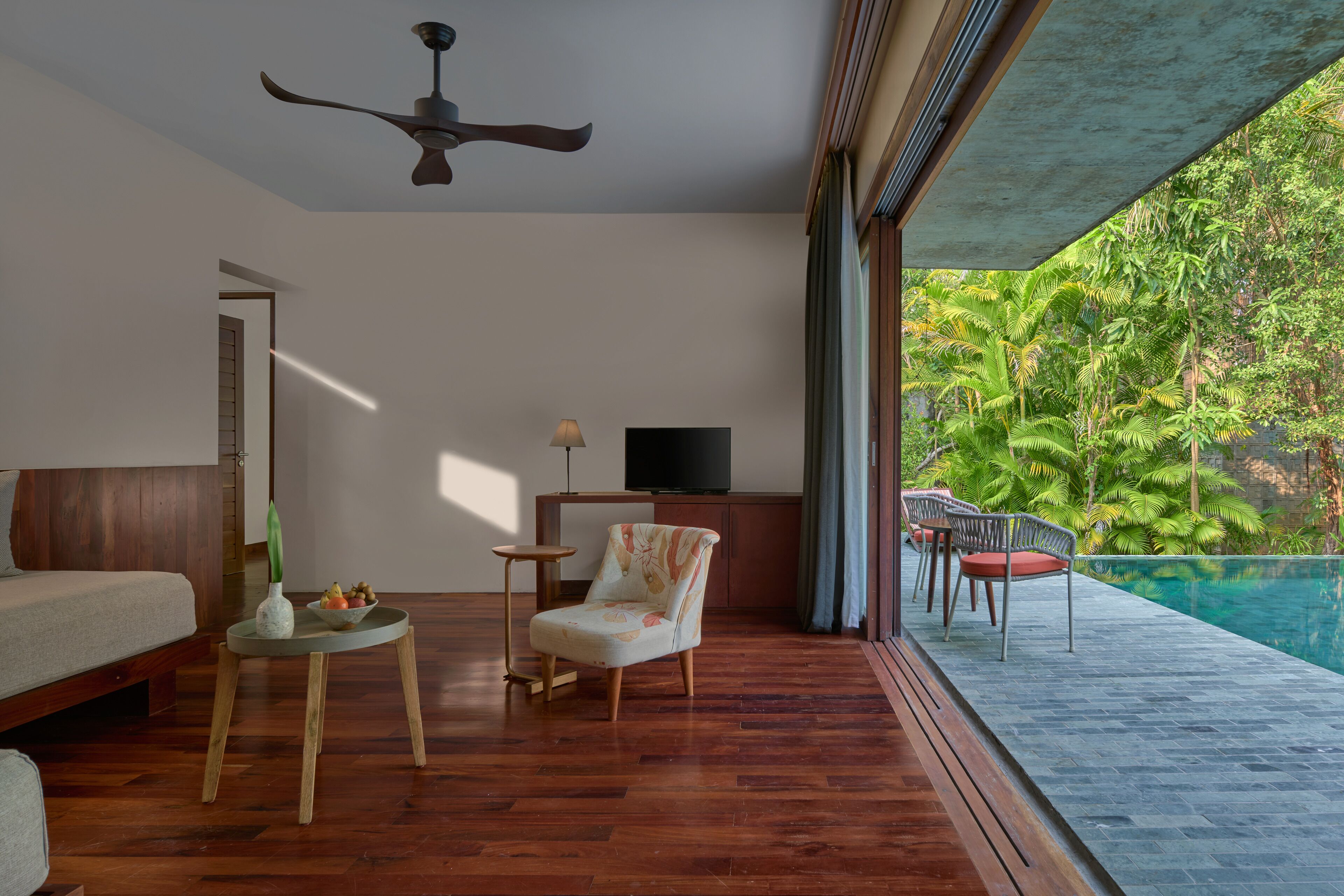 A modern living room with a wooden floor, a sofa, a chair, a small table with a vase and fruit, a television, and a view of a swimming pool and tropical vegetation.