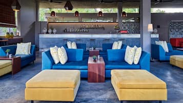 A modern lounge area with blue sofas and yellow ottomans, a bar with a variety of bottles, and a view of greenery outside.