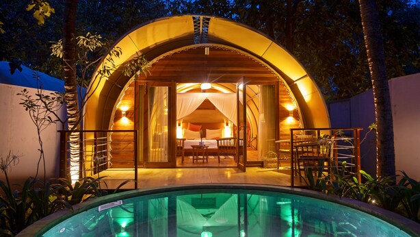 Estructura circular de madera con entrada de vidrio, una piscina al frente y una cama a la vista en su interior.