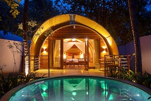 A cozy, circular, wooden structure with a glass entrance, a pool in the foreground, and a bed visible inside.