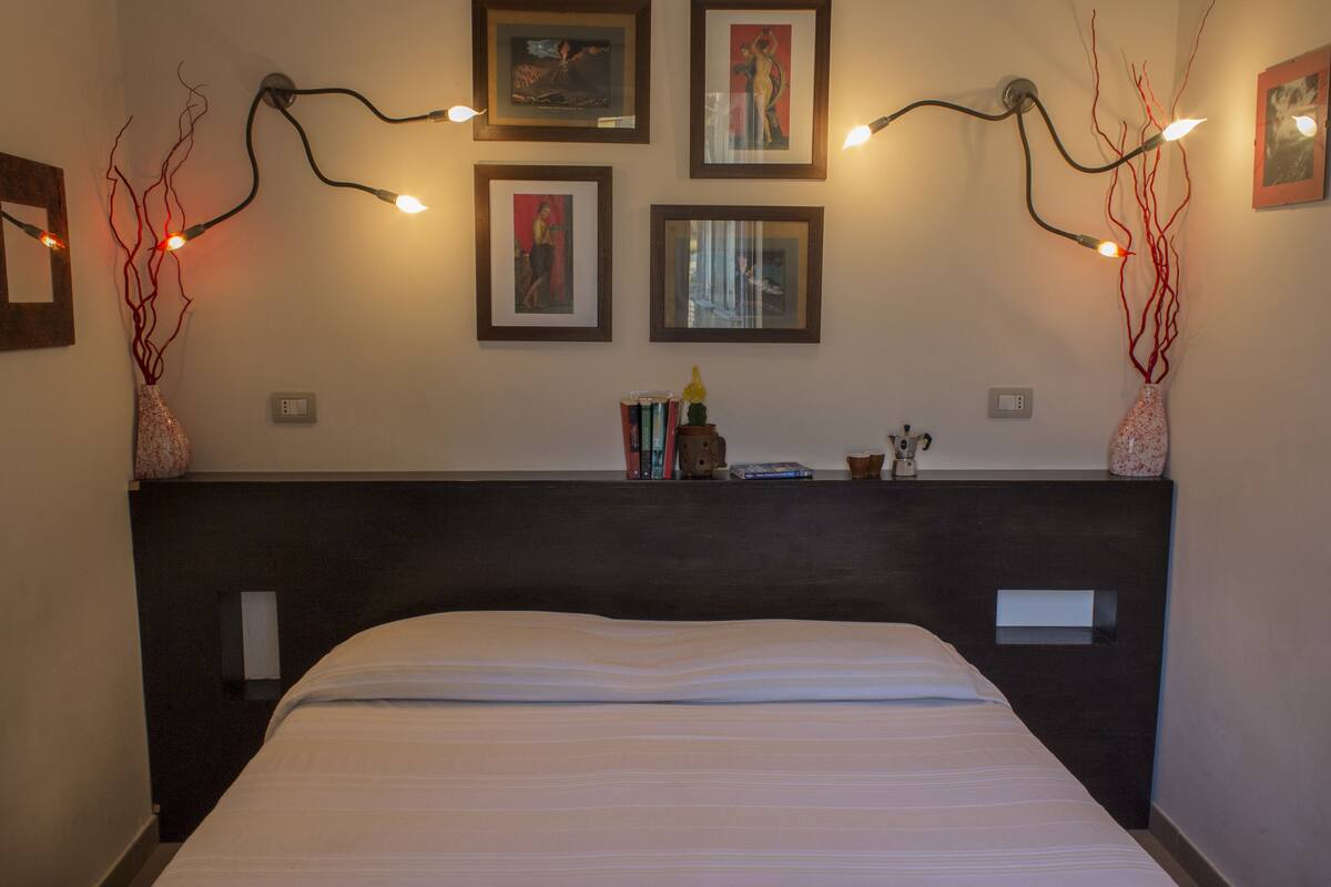 Ein ordentlich bezogenes Bett mit dunklem Kopfteil, ein an der Wand befestigtes Regal mit Büchern und Dekoration sowie gerahmte Kunstwerke darüber.