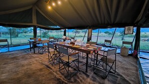 A dining area with a long table set for a meal, surrounded by chairs, under a large tent with a view of the outdoors. - Kananga Special Tented Camp (Serengeti)